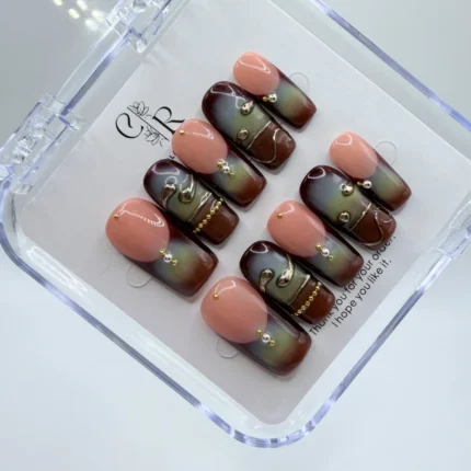 Handmade square press-on nails with brown and green airbrushed aura design, peach French tips, and 3D metallic gold liquid accents in a clear display case.