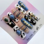 Handmade 3cm square press-on nails featuring blue denim texture, black cow print, and 3D gold bangle charms with blue stones and pearls.