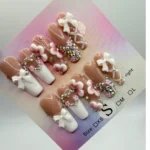 Glossy nude and white press-on nails with 3D bow accents, rhinestones, and pink heart embellishments arranged on a size display card.